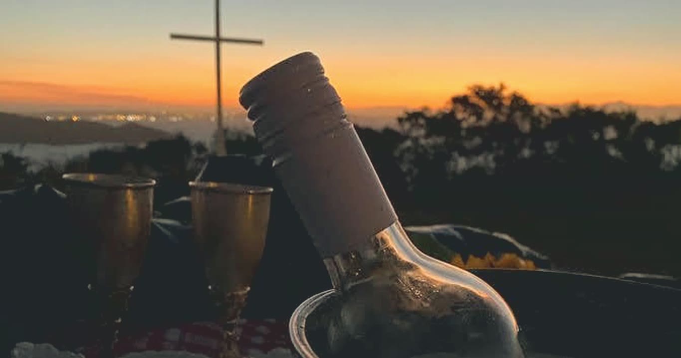Botella y copas frente a la puesta del sol, con una cruz en lo alto del mirador