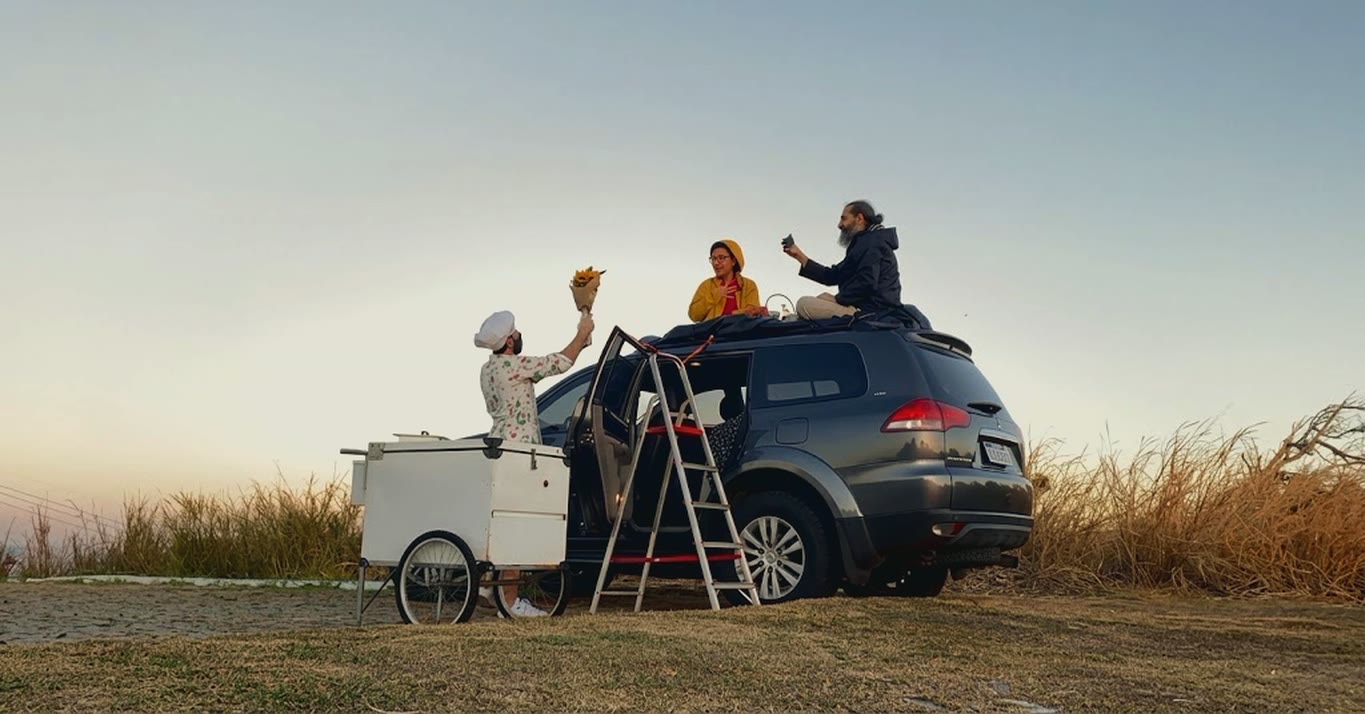 Chef Vitor Jover, beside the ice-cream cart, holds up a bouquet of sunflowers to the couple sitting on the roof of the pickup, at a lookout at sunset