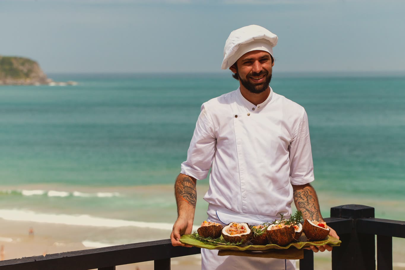 Chef Vitor Jover holding a tray of ceviche served in coconuts, with the sea behind him